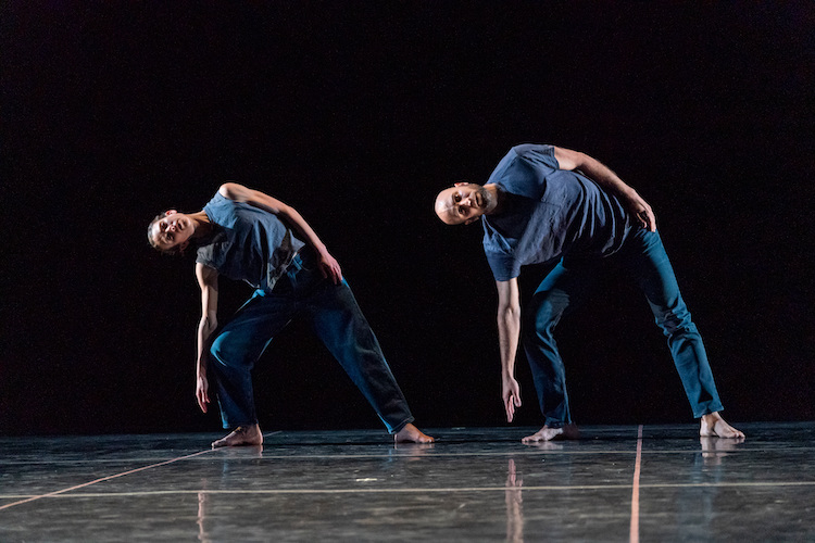 two dancers wearing  blue pants and tshirts lean to one to the right with their right arm extending toward the floor as they stare out at us. Their left arm lies close to their body and the place their left hand on their hip.